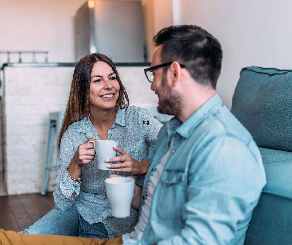 Woman and man sitting on the sofa together. Both are smiling and looking each other ninth eye during their conversation.